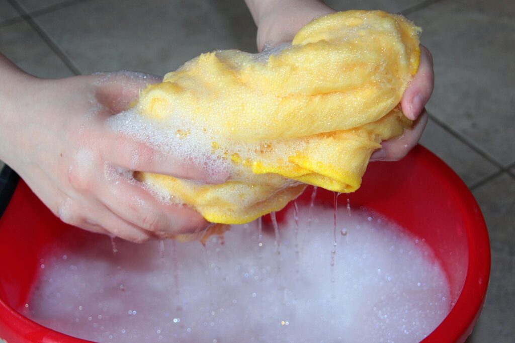 Mãos lavando delicadamente uma blusa de tricô em uma bacia com água e sabão neutro. A imagem transmite cuidado, sem esfregar o tecido, mantendo a integridade dos pontos. 