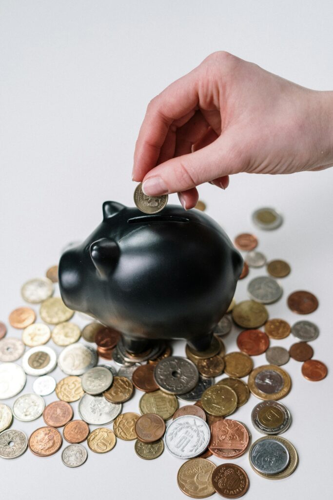 Visão lateral de uma mulher depositando moedas em um cofre, com foco na ação das mãos. A cena simboliza o início da jornada de investimento e a construção da liberdade financeira feminina através de pequenos hábitos diários.