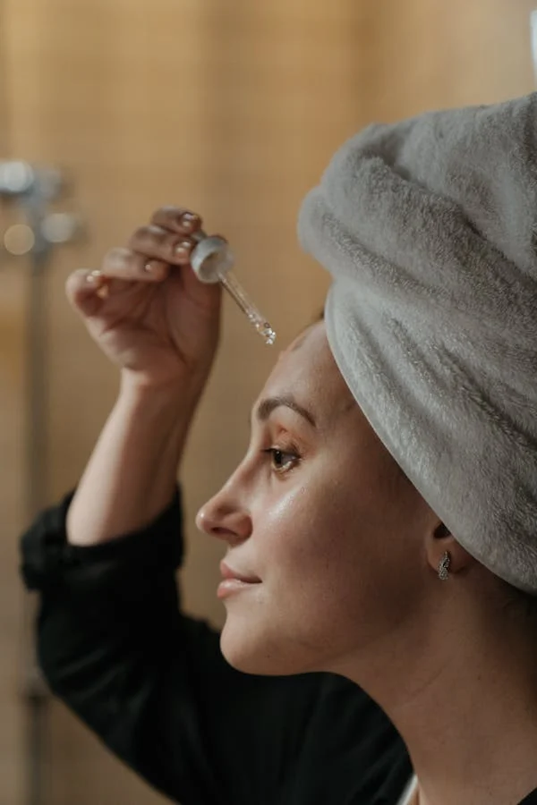 Close-up de uma mulher aplicando um sérum facial. A luz suave da manhã entra pela janela, destacando a textura da pele e criando uma atmosfera de autocuidado e tranquilidade.