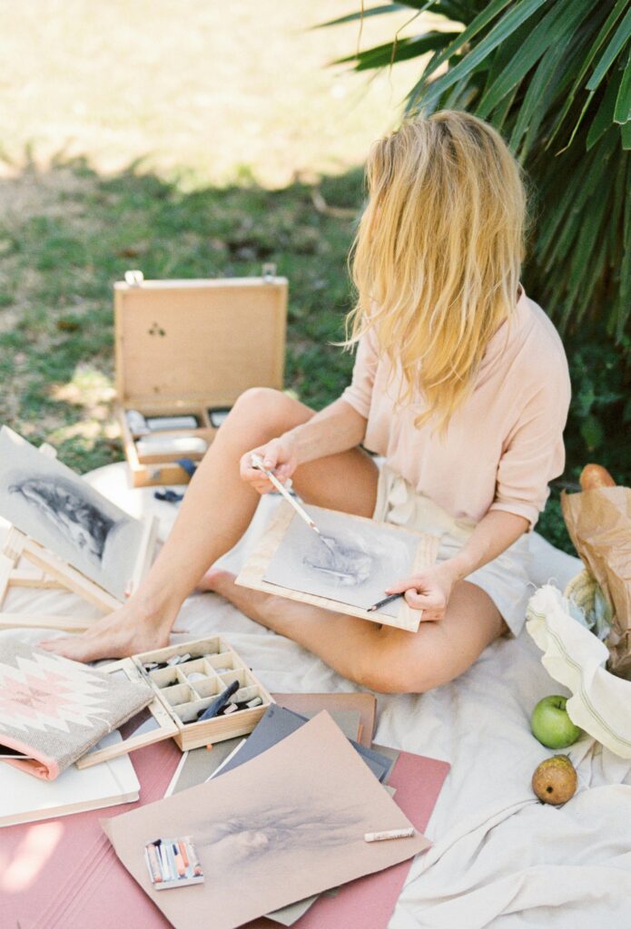Mulher relaxada praticando pintura em uma tela, cercada por plantas e luz natural, simbolizando o prazer e o alívio do estresse através de um hobby