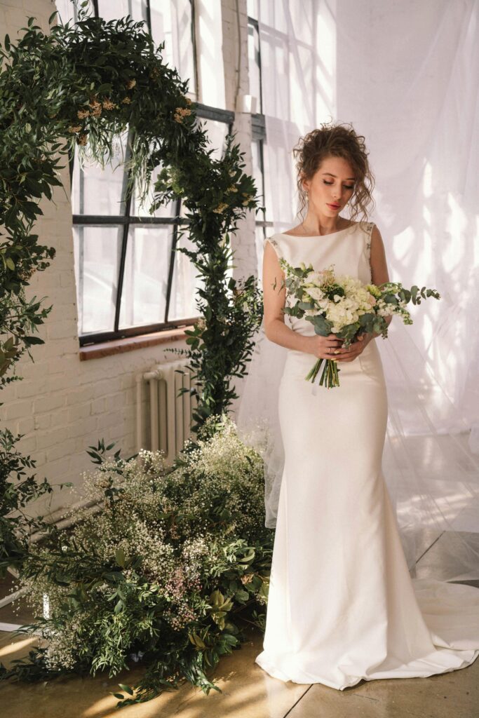 Noiva de perfil usando um vestido branco minimalista de cetim, com corte sereia e decote ombro a ombro, transmitindo elegância e sofisticação em um cenário clean.